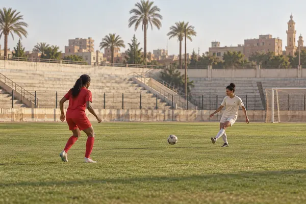 Women footballers passing a ball on a training pitch with palm trees and Egyptian buildings